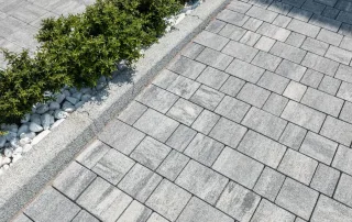 Pavimentazione in blocchi di pietra grigia con bordo in ciottoli e aiuola verde.