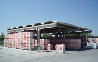 Struttura prefabbricata in cemento con coperture ondulate, utilizzata come area di stoccaggio per materiali da costruzione.