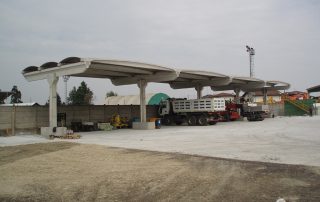 Struttura prefabbricata in cemento con coperture ondulate, utilizzata come area di deposito o parcheggio per automezzi.