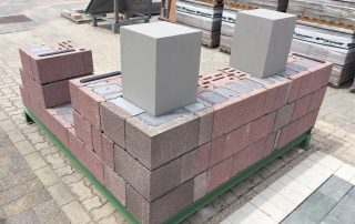 Dimostrazione di muratura con blocchi in laterizio porizzato su una piattaforma espositiva, con colonne grigie e pallet di materiali da costruzione sullo sfondo.