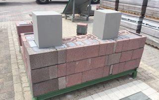 Dimostrazione di muratura con blocchi in laterizio porizzato su una piattaforma espositiva, con colonne grigie e pallet di materiali da costruzione sullo sfondo.