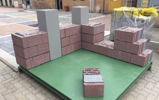 Dimostrazione di muratura con blocchi in laterizio porizzato su una piattaforma espositiva, con colonne grigie e pallet di materiali da costruzione sullo sfondo.