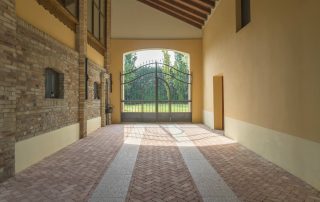 Pavimentazione in mattoni disposti a spina di pesce con fasce in pietra chiara, all'interno di un portico con travi in legno e pareti in mattoni e intonaco.