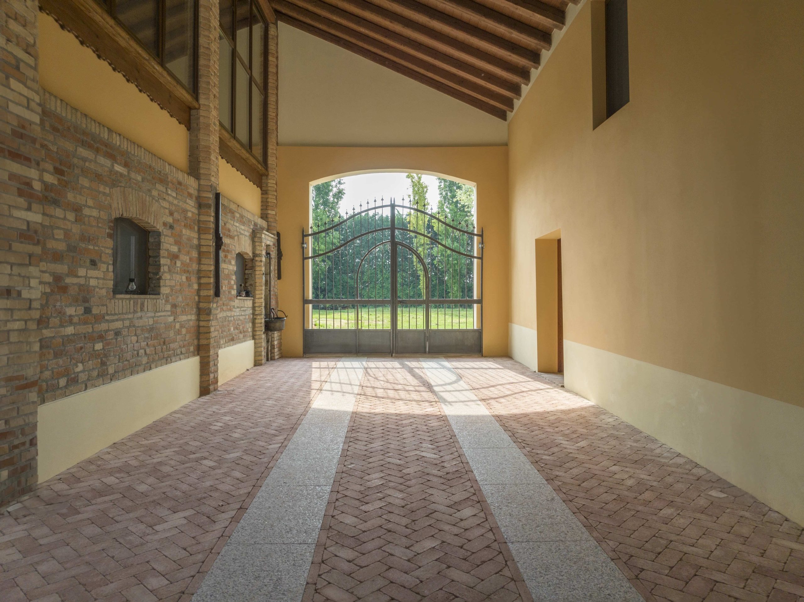 Pavimentazione in mattoni disposti a spina di pesce con fasce in pietra chiara, all'interno di un portico con travi in legno e pareti in mattoni e intonaco.