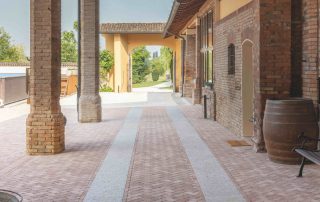 Pavimentazione in mattoni disposti a spina di pesce con fasce in pietra chiara, all'interno di un portico con colonne in mattoni e tetto in legno.