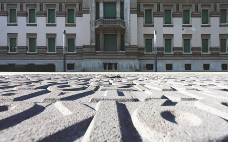 Primo piano di una pavimentazione con incisioni decorative in primo piano, con un edificio storico sullo sfondo caratterizzato da finestre verdi e dettagli architettonici in mattoni e intonaco chiaro.