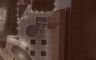 Vista aerea notturna di una piazza con illuminazione a LED integrata, parcheggio adiacente e struttura con tetti in tegole.