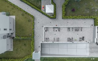 Pavimentazione in masselli grigi con aree verdi delimitate da siepi, vista dall'alto. Spazi organizzati con rastrelliere per biciclette e un gazebo bianco.