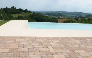Pavimentazione in autobloccanti dal design rustico con finitura anticata, posata attorno a una piscina a sfioro con vista panoramica.