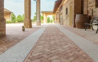 Pavimentazione in mattoni disposti a spina di pesce con fasce in pietra chiara, all'interno di un portico con colonne in mattoni e tetto in legno.