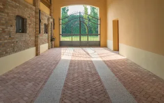 Pavimentazione in mattoni disposti a spina di pesce con fasce in pietra chiara, all'interno di un portico con travi in legno e pareti in mattoni e intonaco.