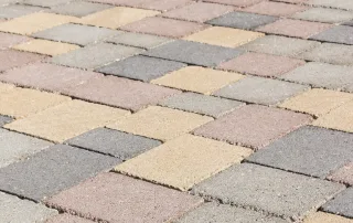 Pavimentazione in blocchi quadrati con tonalità miste di grigio, beige e marrone, disposti in un pattern regolare.