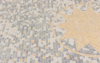 Superficie pavimentata con piccoli blocchi di pietra disposti in un motivo regolare. Al centro, un motivo a stella giallo ocra circondato da un cerchio, contrastante con il resto della pavimentazione in tonalità grigie e beige.