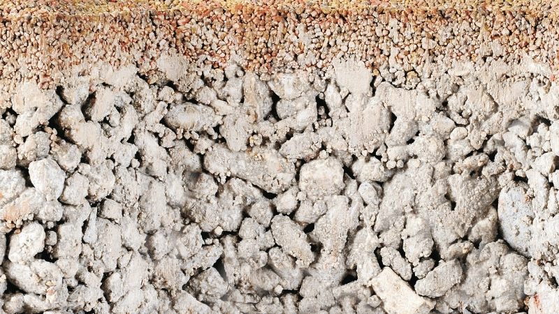 Sezione di pavimentazione filtrante composta da uno strato superficiale di ghiaia fine di colore marrone e da uno strato inferiore in calcestruzzo poroso grigio.