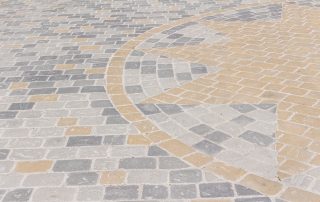 Pavimentazione in pietra con un motivo geometrico composto da lastre rettangolari e quadrate in diverse tonalità di grigio e ocra.
