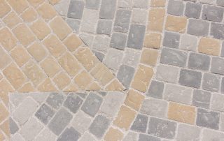 Pavimentazione in pietra con un motivo geometrico composto da lastre rettangolari e quadrate in diverse tonalità di grigio e ocra.