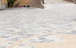 Pavimentazione in pietra con un motivo geometrico composto da lastre rettangolari e quadrate in diverse tonalità di grigio e ocra.
