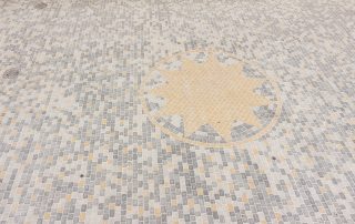 Superficie pavimentata con piccoli blocchi di pietra disposti in un motivo regolare. Al centro, un motivo a stella giallo ocra circondato da un cerchio, contrastante con il resto della pavimentazione in tonalità grigie e beige.