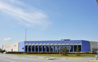 Facciata di un capannone industriale prefabbricato con pannelli di cemento e rivestimenti metallici blu con colonne blu e struttura in acciaio con ampie superfici vetrate.