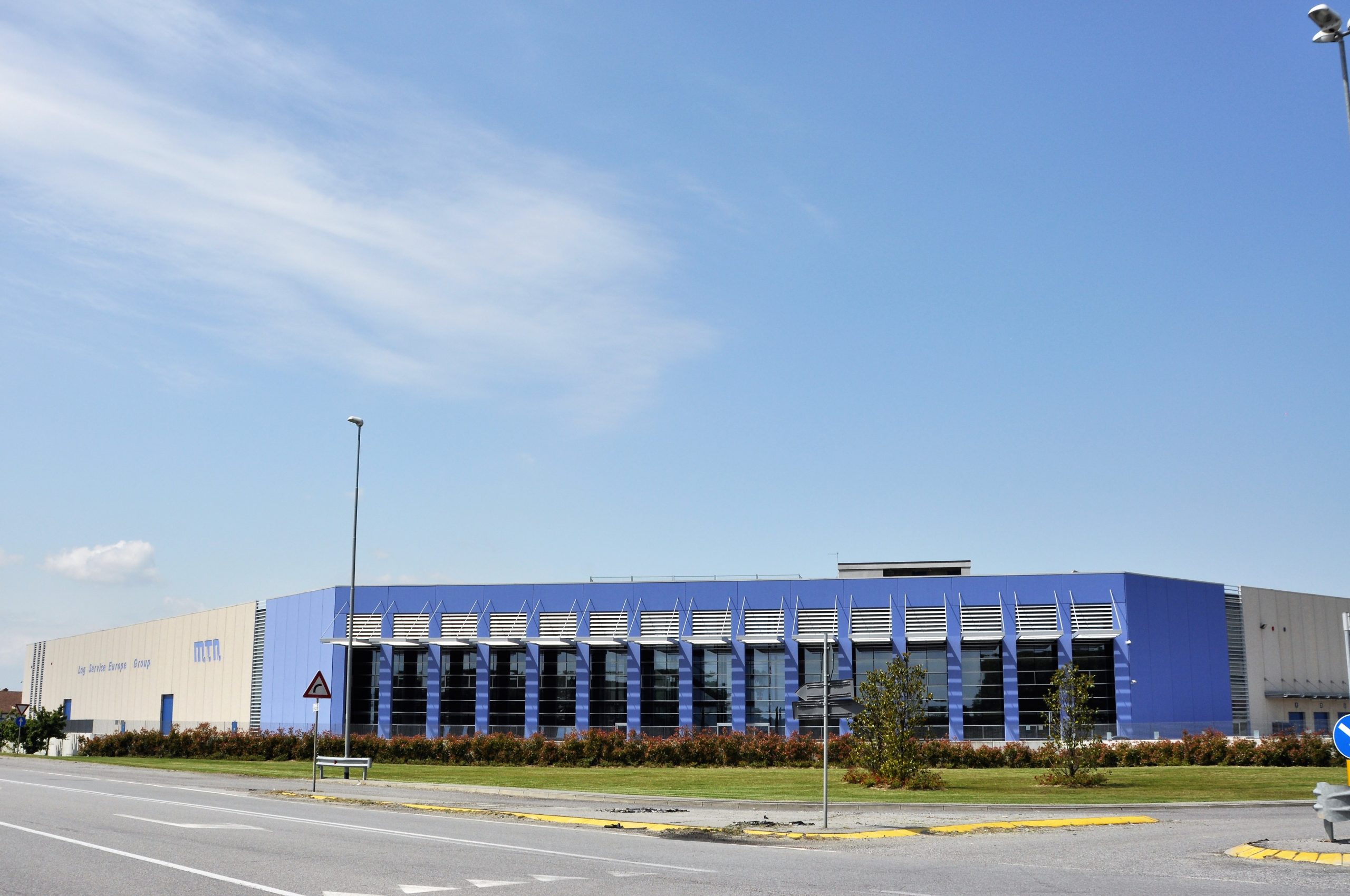 Facciata di un capannone industriale prefabbricato con pannelli di cemento e rivestimenti metallici blu con colonne blu e struttura in acciaio con ampie superfici vetrate.
