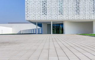 Pavimentazione in lastre rettangolari chiare con linee sottili di separazione, davanti a un edificio moderno bianco con pannelli decorati da motivi di farfalle.