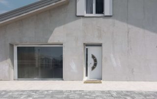 Pavimentazione in pietra grigia in primo piano, estendendosi fino all'ingresso di una casa moderna con pareti in cemento chiaro e finestre bianche.