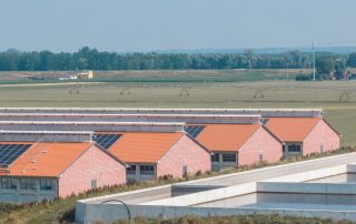 Complesso di edifici agricoli con tetti a doppia falda ricoperti da pannelli solari. Le strutture in mattoni rossi sono immerse in un paesaggio rurale, con vasche di raccolta dei liquami in primo piano.