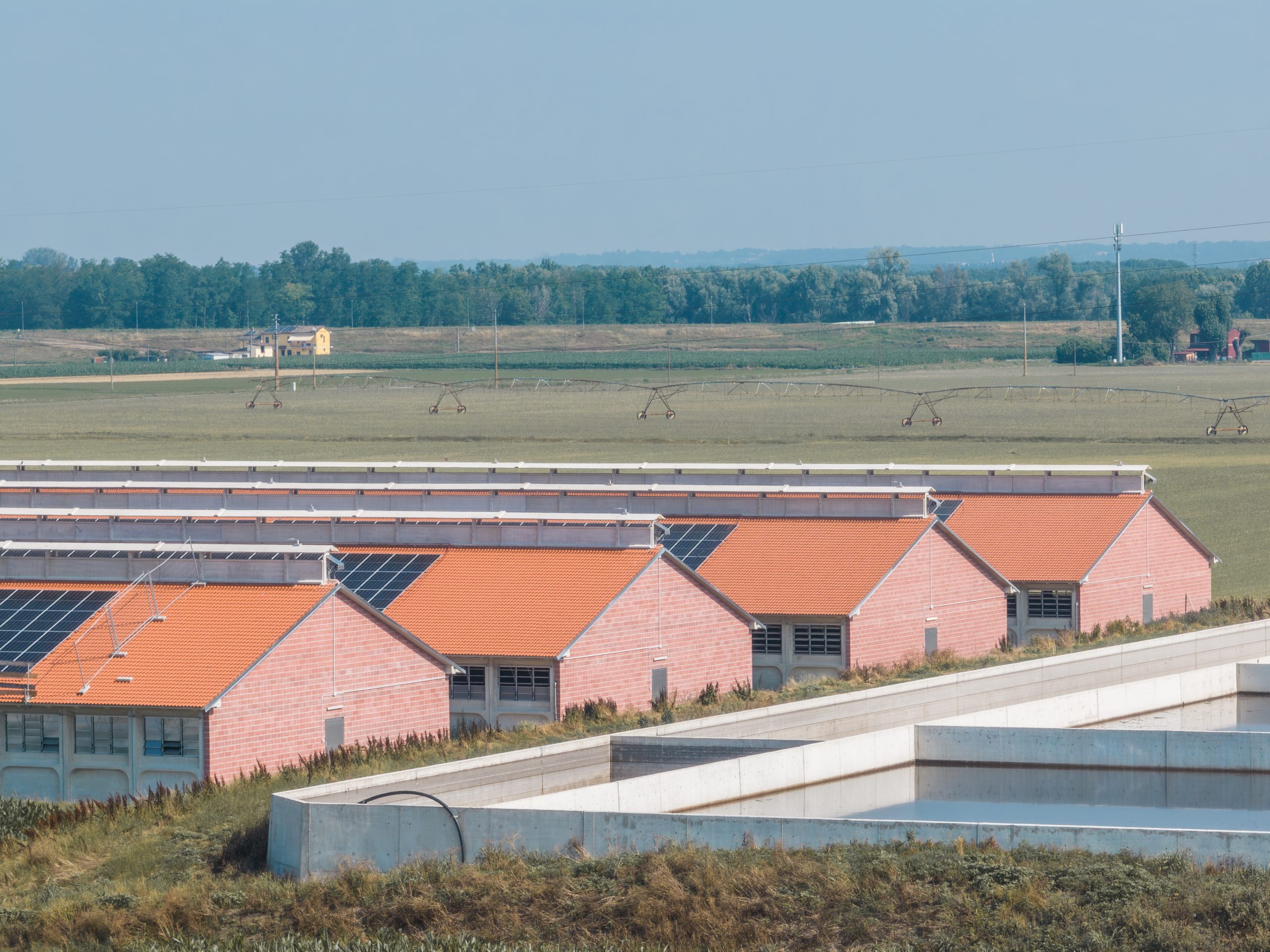 Complesso di edifici agricoli con tetti a doppia falda ricoperti da pannelli solari. Le strutture in mattoni rossi sono immerse in un paesaggio rurale, con vasche di raccolta dei liquami in primo piano.