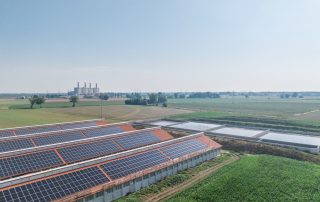 Complesso di edifici agricoli con tetti a doppia falda ricoperti da pannelli solari. Le strutture in mattoni rossi sono immerse in un paesaggio rurale, con vasche di raccolta dei liquami.