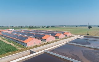Complesso di edifici agricoli con tetti a doppia falda ricoperti da pannelli solari. Le strutture in mattoni rossi sono immerse in un paesaggio rurale, con vasche di raccolta dei liquami in primo piano.