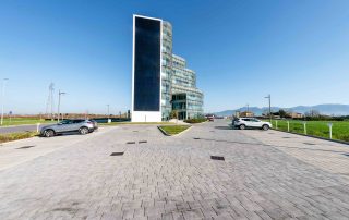 Ampia pavimentazione in blocchi rettangolari effetto pietra con tonalità neutre, in primo piano, che conduce a un moderno edificio in vetro e acciaio con forme curve e facciata trasparente.