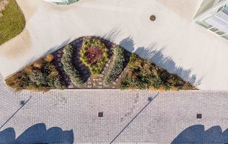 Vista dall'alto di una pavimentazione in pietra con tonalità neutre, affiancata da un'area verde curata con aiuole simmetriche.