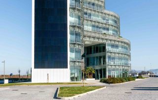 Ampia pavimentazione in blocchi rettangolari effetto pietra con tonalità neutre, in primo piano, che conduce a un moderno edificio in vetro e acciaio con forme curve e facciata trasparente.