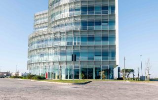 Ampia pavimentazione in blocchi rettangolari effetto pietra con tonalità neutre, in primo piano, che conduce a un moderno edificio in vetro e acciaio con forme curve e facciata trasparente.