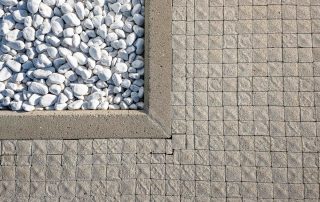 Pavimentazione in cubetti di pietra con finitura ruvida, affiancata da un'aiuola bordata in cemento contenente ciottoli bianchi decorativi.