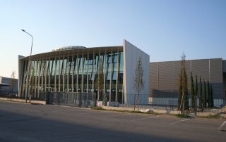 Edificio moderno con ampia facciata in vetro e struttura inclinata in metallo, affiancato da una sezione con pannelli metallici.