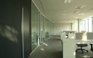 Ufficio moderno e luminoso con open space, arredato con postazioni di lavoro bianche e pareti divisorie in vetro.
