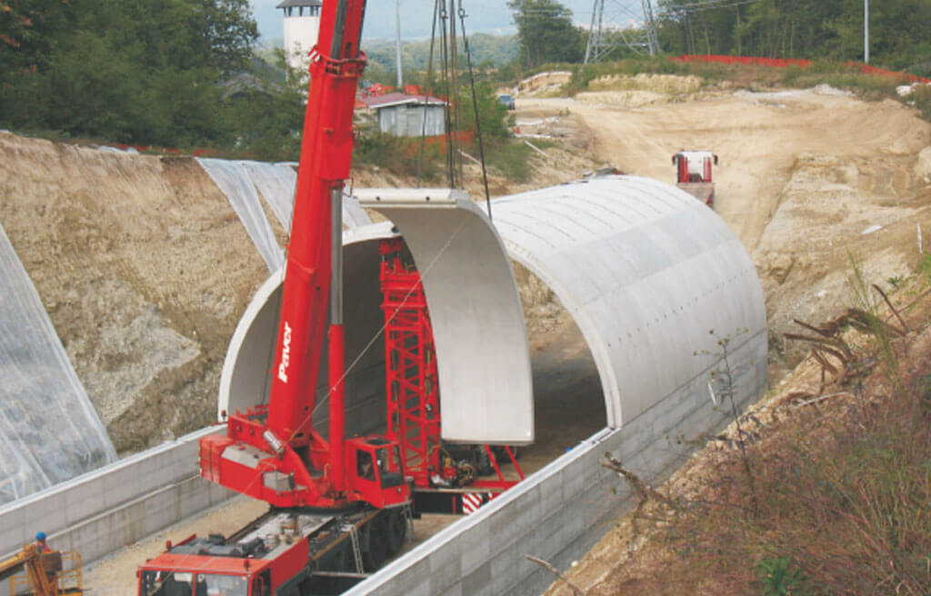 Costruzione di un tunnel con una gru rossa che solleva un grande segmento prefabbricato in cemento.