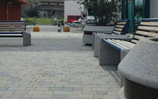 Pavimentazione in blocchi di pietra rettangolari di varie tonalità di grigio, con panchine e fioriere in cemento.
