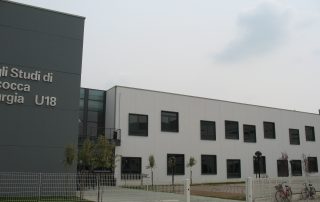 Edificio prefabbricato dell'Università degli Studi di Milano Bicocca, Medicina e Chirurgia U18, con facciata moderna in pannelli grigi e bianchi, finestre nere incassate e recinzione metallica.