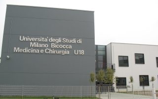 Edificio prefabbricato dell'Università degli Studi di Milano Bicocca, Medicina e Chirurgia U18, con facciata moderna in pannelli grigi e bianchi, finestre nere incassate e recinzione metallica.