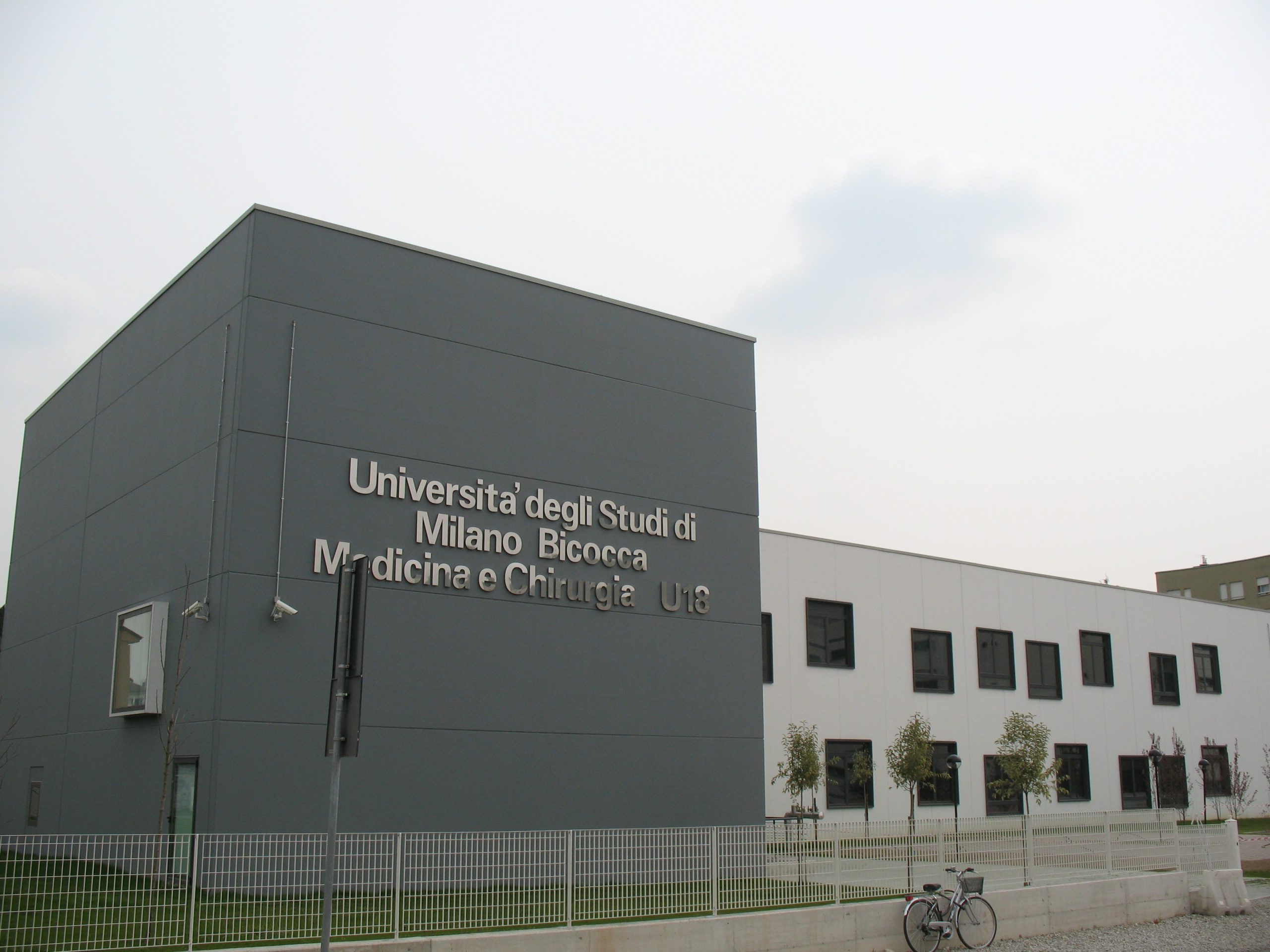 Edificio prefabbricato dell'Università degli Studi di Milano Bicocca, Medicina e Chirurgia U18, con facciata moderna in pannelli grigi e bianchi, finestre nere incassate e recinzione metallica.