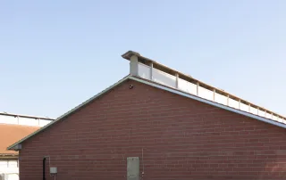 Vista laterale di una porcilaia con parete in mattoni rossi e tetto spiovente. La struttura presenta un sistema di ventilazione sopraelevato sul colmo del tetto.