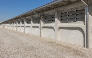 Vista esterna di una porcilaia con struttura in cemento prefabbricato. La facciata presenta una serie di finestre a lamelle regolabili in metallo, collegate a un sistema di ventilazione automatizzato per la gestione del flusso d'aria. Sono visibili anche canalizzazioni per il drenaggio dell'acqua piovana lungo il tetto.