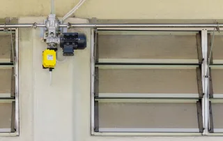 Dettaglio di un sistema di ventilazione automatizzato su una porcilaia. L'immagine mostra una serie di finestre a lamelle regolabili in metallo e policarbonato, collegate a un motore elettrico con un attuatore giallo e un sistema di aste e leve in acciaio inox. Questo sistema consente il controllo automatico dell'apertura e chiusura delle prese d'aria, regolando la ventilazione all'interno dell'edificio per garantire condizioni ottimali per gli animali.