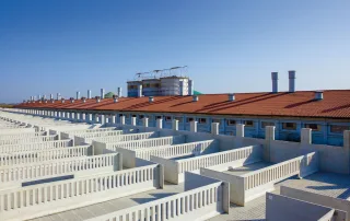 Vista panoramica di una grande porcilaia con numerosi recinti in cemento e pavimenti grigliati, progettati per la gestione efficiente degli allevamenti suini. Il tetto in tegole rosse è dotato di camini di aerazione. Sullo sfondo, si notano grandi silos e strutture per la gestione dell'azienda agricola.