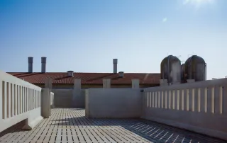 Struttura di una porcilaia con pavimento grigliato in cemento. Sullo sfondo si distinguono silos metallici e camini di aerazione sul tetto in tegole rosse.