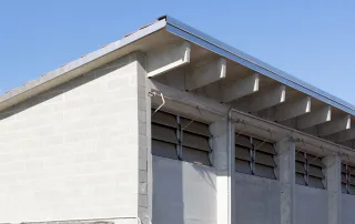 Dettaglio angolare di una porcilaia con pareti in blocchi di cemento e tetto in tegole, rifinito con una lamiera metallica. Le finestre a lamelle regolabili garantiscono la ventilazione.