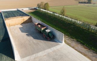 Vista aerea di un'area di stoccaggio agricola con silos in cemento armato prefabbricato. Un trattore con rimorchio è posizionato all'interno di uno dei comparti, parzialmente riempito di foraggio o insilato. A sinistra, un altro comparto è coperto con un telo di protezione.