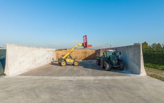 Vista di un silo agricolo in cemento armato prefabbricato, parzialmente riempito con insilato. Un sollevatore telescopico Dieci con attrezzatura Trioliet TU 180 XL è in azione per il caricamento del materiale in un rimorchio trainato da un trattore Fendt.